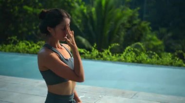 Çekici Avrupalı esmer spor kızı doğada yoga yapıyor. Havuzun kenarında yeşil bir yağmur ormanı var ve derin derin nefes alıyor..