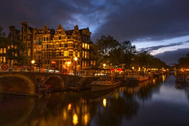 Hollanda 'da. Amsterdam Kanalı 'nda bulutlu bir akşam. Tekneler ve yüzen evler demirli. Geleneksel evlerin sularında yansıma ve bir köprü