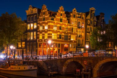 Hollanda 'da. Amsterdam Kanalı 'nda bir gece. Işıklı otantik Hollanda evi. Köprüdeki arabalardan hafif izler. Demir atmış eski bir tekne