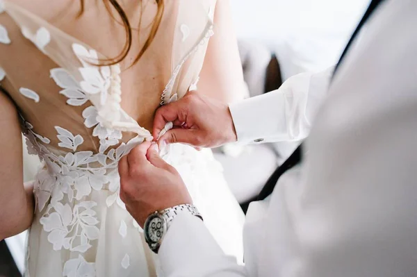ネクタイと時計のついた白いシャツの男は ドレスのコルセットにボタンを固定します 結婚式のドレスで花嫁は部屋に立ってレース — ストック写真