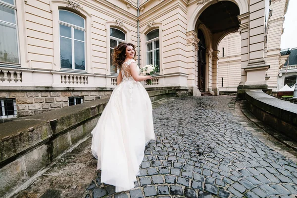 古い石畳の上を旧市街を歩くウェディングドレスの花嫁 結婚式の花束を持つ女性は宮殿に戻ります — ストック写真