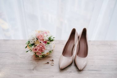 Pembe çiçeklerin, güllerin ve yeşilliğin gelininin düğün buketi. Renkli klasik bej ayakkabılar, küpe ve yüzük pastel arka planda duruyor. Gelin aksesuarları. Kapatın. Düz yat. en üstteki görünüm.