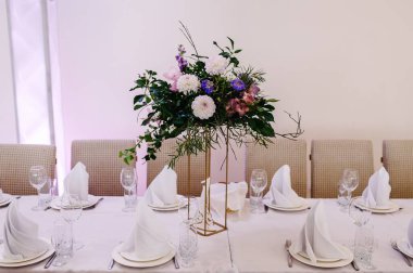 Masa masa örtüsüyle örtülmüş. Tabaklar ve çatal bıçaklarla servis edilmiş. Düğün salonundaki çiçek ve yeşillik kompozisyonu ile süslenmiş şölen masası..