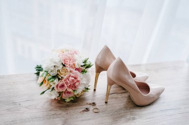 Pembe çiçeklerin, güllerin ve yeşilliğin gelininin düğün buketi, süslü klasik bej ayakkabılar, küpe ve yüzük ahşap arka planda yatıyor. Gelin aksesuarları. Kapatın. Yan görünüm.