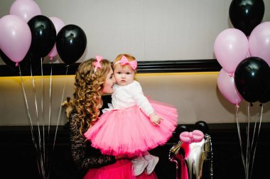 Siyah ve pembe balonların arka planında pembe tüllü bir etek giyen anne ve kız. Küçük bir kız bebek prensesin yanağından öpüyor. Doğum günü kutlaması 1 yıldır..