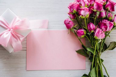 Pembe küçük güller, hediye kutusu, beyaz ahşap arka planda kartpostal. Doğum günün kutlu olsun. Bayram tebrik kartı, sevgililer günü, kadınlar, anneler günü, paskalya. Mesaj için yer açın. Üst manzara, düz uzanma.