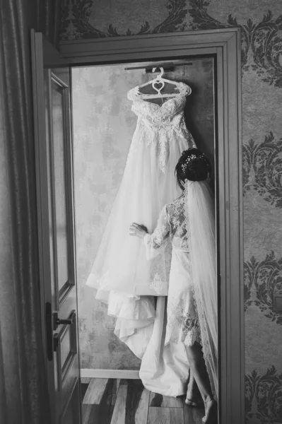 Bride holding a dress in room, girl in sexy night dress at home. Preparing for a wedding in the morning. Back view. Black and white photo.