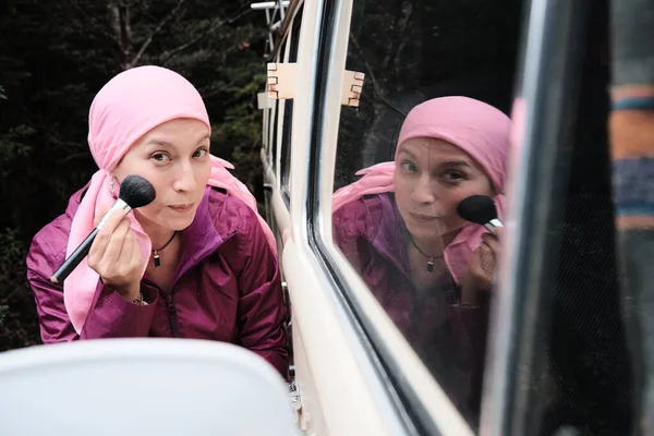 A young cancer survivor wearing a pink scarf puts on makeup and smiles while looking into a car mirror