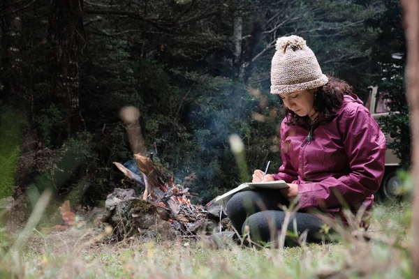 Beautiful traveler writing in a notebook while resting near a campfire in the forest