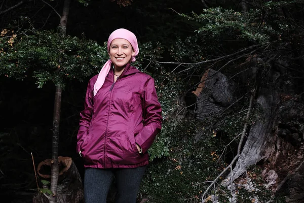 Smiling female cancer survivor enjoys a day in the countryside. She wears a pink scarf and purple jacket.