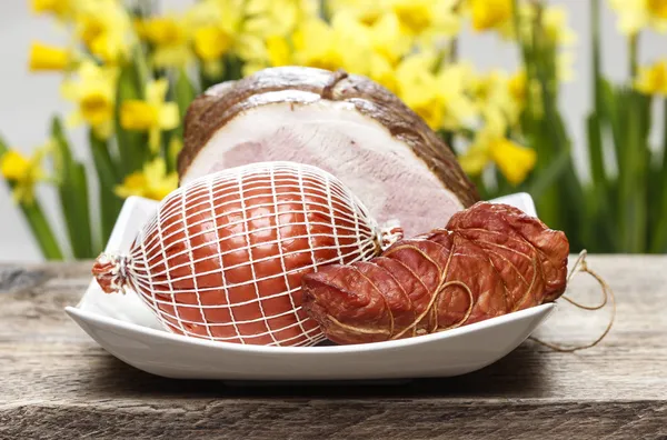 Smoked ham on wooden table. Yellow daffodils in the background. — Stock Photo, Image