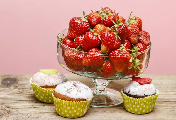 Glass bowl of strawberries and colorful muffins on wooden table. — Stock Photo, Image