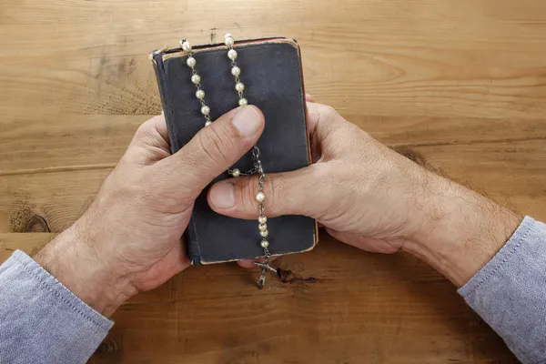 Hands with rosary over old Holy Bible. Wooden background. — Stock Photo, Image