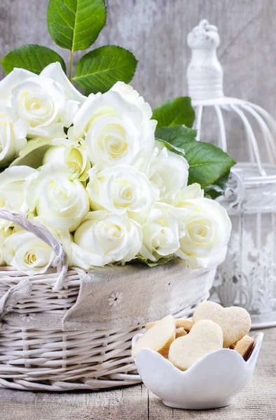 Wicker basket of white roses and bowl of cookies on rustic woode — Stock Photo, Image