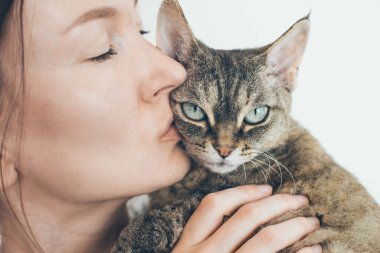Güzel gözlü mutlu kedi portresi ve sarılan genç bir kadın. Yakışıklı dişi sarılıyor ve sevimli Devon Rex yavrusunu öpüyor. Evcil hayvanlar. Kitty dikkat çekmeyi ve mırıldanmayı sever.