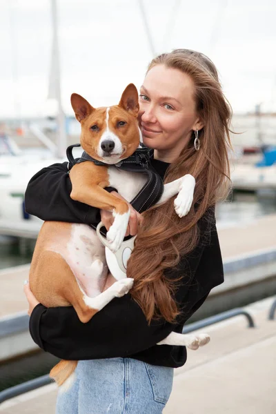 Portrait of happy young female with her basenji male dog. Woman is holding and hugging her cute doggy while walking outdoors. Positive emotions. Dog is feeling relaxed with the owner.