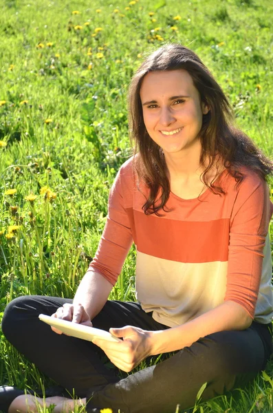 Menina morena atraente bonita está sentada em um prado com tablet — Fotografia de Stock