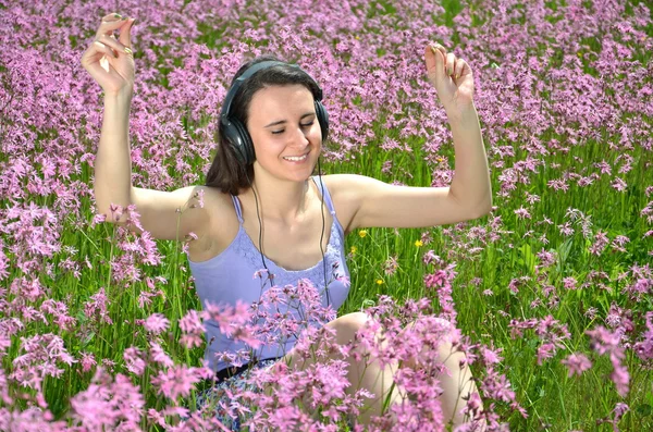 Menina morena atraente bonita ouvindo música com fones de ouvido no prado lindo cheio de flores selvagens — Fotografia de Stock