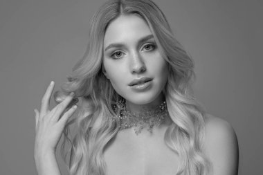 Gri arka planda uzun kıvırcık saçlı güzel bir sarışının portresi boynunda küçük taşlarla süslenmiş. Siyah beyaz fotoğraf.