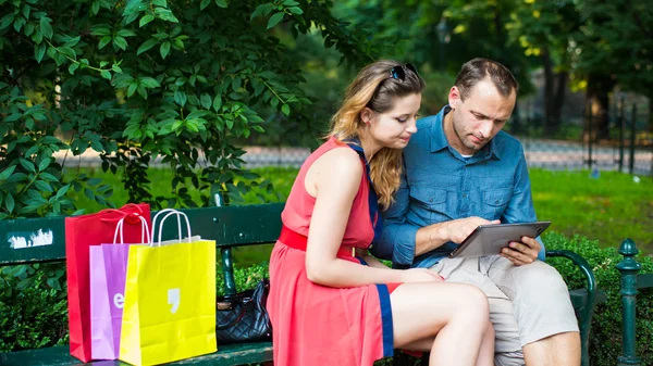 Пара сидящих на скамейке с сумками и планшетом — стоковое фото