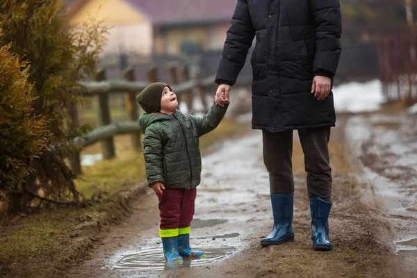 Padre e figlio che camminano all'aria aperta in stivali di gomma sulle pozzanghere e fango dopo la pioggia. Bambino che tiene la mano di un uomo — Foto Stock