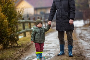 Baba ve oğul temiz havada, su birikintilerinde lastik botlarla yağmurdan sonra çamurda yürüyorlar. Küçük çocuk bir adamın elini tutuyor.