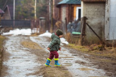 Koruyucu lastik botları ve çamur birikintisinde zıplayan yağmurluk giymiş küçük bir çocuk. Mutlu çocuk yağmur üstüne su birikintisinde oynarken eğleniyor..