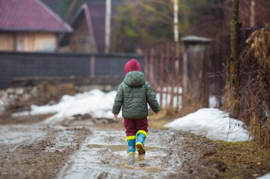 Koruyucu lastik botları ve çamur birikintisinde zıplayan yağmurluk giymiş küçük bir çocuk. Mutlu çocuk yağmur üstüne su birikintisinde oynarken eğleniyor..