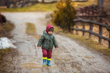 Koruyucu lastik botları ve çamur birikintisinde zıplayan yağmurluk giymiş küçük bir çocuk. Mutlu çocuk yağmur üstüne su birikintisinde oynarken eğleniyor..