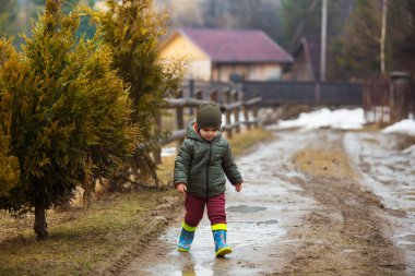 Koruyucu lastik botları ve çamur birikintisinde zıplayan yağmurluk giymiş küçük bir çocuk. Mutlu çocuk yağmur üstüne su birikintisinde oynarken eğleniyor.