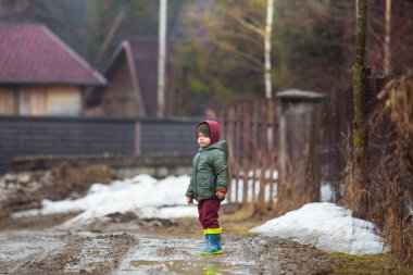Koruyucu lastik botları ve çamur birikintisinde zıplayan yağmurluk giymiş küçük bir çocuk. Mutlu çocuk yağmur üstüne su birikintisinde oynarken eğleniyor.