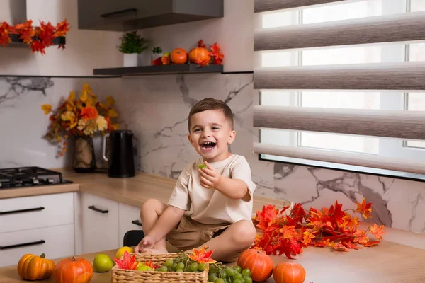 Smiling cute sitting on kitchen table with fruits, autumn leaves and pumpkin. Autumn decorated kitchen. Holidays concept