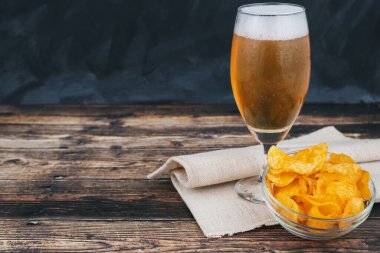 Koyu ahşap arka planda bir kase cipsin yanında taze çekilmiş hafif bira bardağı.