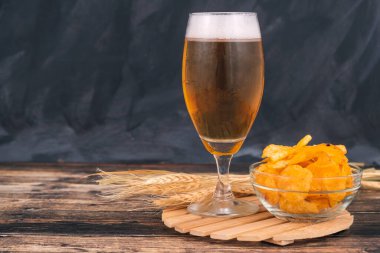 Koyu ahşap arka planda bir kase cipsin yanında taze çekilmiş hafif bira bardağı.