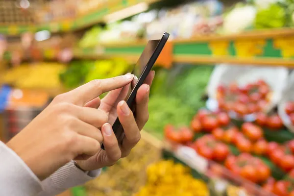 Mujer usando el teléfono móvil mientras va de compras en el supermercado Imágenes de stock libres de derechos