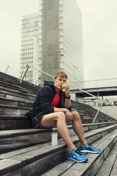 Şehir koşucusu idmandan sonra yeşil bir elmayı ısıtıyor. Şehir arkaplanı