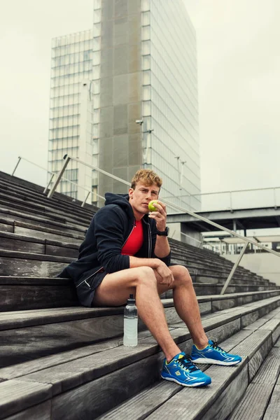Şehir koşucusu idmandan sonra yeşil bir elmayı ısıtıyor. Şehir arkaplanı