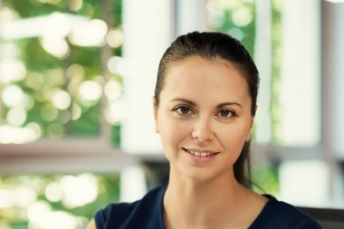 İş yerindeki doğal genç kadın portresi. Kameraya bakıyorum. Büyük pencereler ve ağaçlar