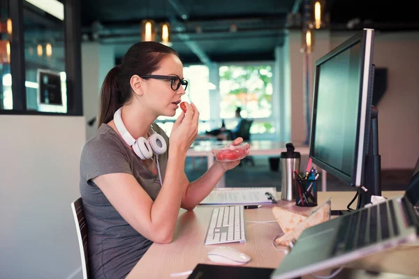 現代のオフィスでコンピュータ画面の前でトマトを食べるカジュアルな若いビジネス女性 — ストック写真