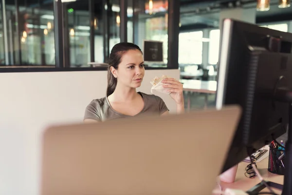ランチタイムには 現代的なビジネスオフィススペースでコンピュータ画面の前でサンドイッチを食べるカジュアルな若い女性 — ストック写真
