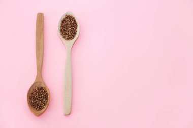 Pembe arka planda, tahta bir kaşıkta, doğal kahverengi karabuğday taneleri.