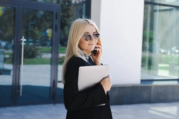 A young woman in a suit is talking on the phone and holding a laptop in her hands on the street