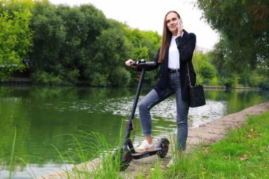 Genç bir kadın telefonda parkta elektrikli bir scooterın yanında konuşuyor.