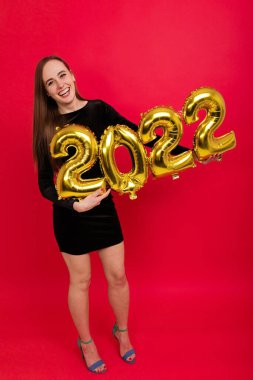 Siyah elbiseli ve topuklu mutlu bir esmer, 2022 rakamlarını kırmızı arka planda tutar.