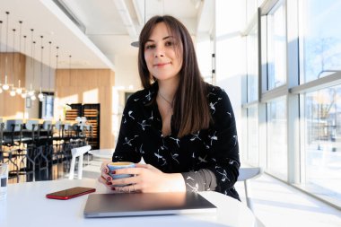 A brunette in a black dress drinks coffee during breaks from work in a cafe