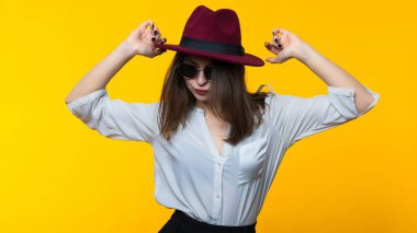 Sarı arka planda dans eden şapkalı ve gözlüklü genç bir kadın.