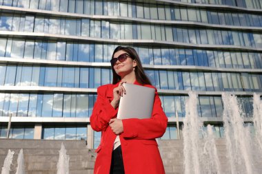 Ceketli bir iş kadını, bir ofis binasının arka planında dizüstü bilgisayarla poz veriyor.