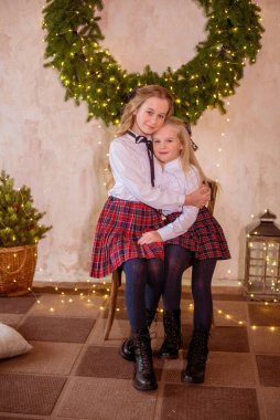 Sarışın, kıvırcık saçlı, klasik okul kıyafetleriyle gişelere yasaklı güzel kız kardeşler duvarda klasik bir iç mekan olan çam iğnelerinden oluşan büyük bir Noel çelenginin yanında. Noel havası 