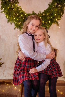 Sarışın, kıvırcık saçlı, klasik okul kıyafetleriyle gişelere yasaklı güzel kız kardeşler duvarda klasik bir iç mekan olan çam iğnelerinden oluşan büyük bir Noel çelenginin yanında. Noel havası 