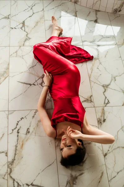 一位年轻貌美的姑娘 一头乌黑的长发 晚上化着妆 穿着一件长长的红色晚礼服 躺在大理石地板上 美与时尚 — 图库照片